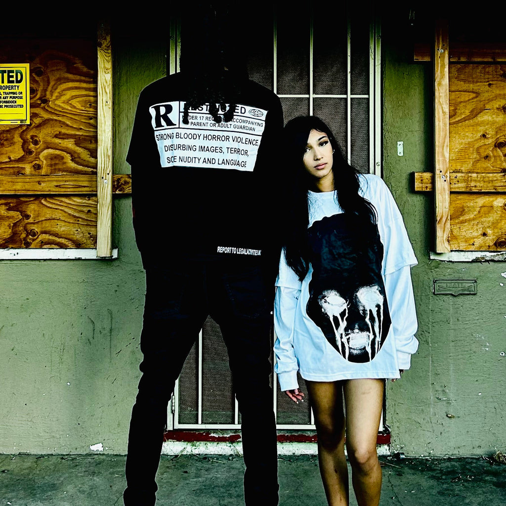 Two models posing in front of a boarded-up motel room. One model is shown from the back wearing a black hoodie with a large white graphic print on the back, and the other model is wearing a light blue oversized sweatshirt with a large black skull graphic on the front.