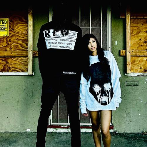 Two models posing in front of a boarded-up motel room. One model is shown from the back wearing a black hoodie with a large white graphic print on the back, and the other model is wearing a light blue oversized sweatshirt with a large black skull graphic on the front.